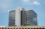 A tall modern building with reflective windows and 'BCH' sign on the side, against a clear blue sky. The foreground shows part of a roof with clay tiles and some greenery.
