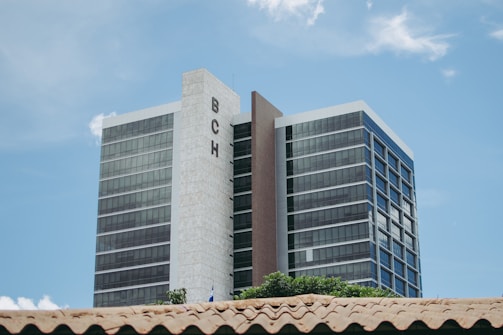 A tall modern building with reflective windows and 'BCH' sign on the side, against a clear blue sky. The foreground shows part of a roof with clay tiles and some greenery.