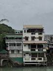 A multi-story building stands alongside a river with a rustic appearance. The structure has several balconies and is adorned with plants. There's a sign on the ground floor that reads 'La Vista Pagsanjan Resort.' The overall setting is serene, with a cloudy sky and greenery surrounding the area.