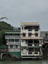 A multi-story building stands alongside a river with a rustic appearance. The structure has several balconies and is adorned with plants. There's a sign on the ground floor that reads 'La Vista Pagsanjan Resort.' The overall setting is serene, with a cloudy sky and greenery surrounding the area.