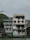 A multi-story building stands alongside a river with a rustic appearance. The structure has several balconies and is adorned with plants. There's a sign on the ground floor that reads 'La Vista Pagsanjan Resort.' The overall setting is serene, with a cloudy sky and greenery surrounding the area.