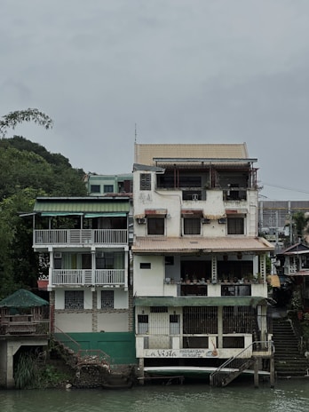 A multi-story building stands alongside a river with a rustic appearance. The structure has several balconies and is adorned with plants. There's a sign on the ground floor that reads 'La Vista Pagsanjan Resort.' The overall setting is serene, with a cloudy sky and greenery surrounding the area.