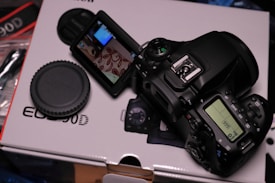 A digital DSLR camera is placed on its box, displaying a flip-out screen which shows part of a patterned surface. The camera is positioned on its side, revealing various control buttons and a backlit display screen with the number 999. A detachable lens cap is present next to the camera.