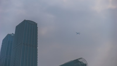 A skyline with tall skyscrapers under a cloudy sky, featuring a commercial airplane flying in the distance.