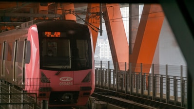 A pink metro train moves along tracks on a bridge with prominent orange structural beams. The front of the train displays the destination as 'To Beibei' in red text. The surrounding environment is industrial with metal fencing and overhead wires.