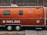 A vintage Airstream trailer with an orange exterior is parked in front of an industrial building with large grid-like windows. The trailer features the word 'INKLING' in bold white letters and some additional text, suggesting it is associated with a brand or company. A website URL is also visible on the trailer.