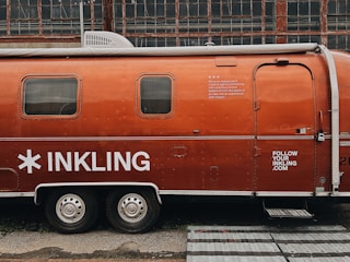 A vintage Airstream trailer with an orange exterior is parked in front of an industrial building with large grid-like windows. The trailer features the word 'INKLING' in bold white letters and some additional text, suggesting it is associated with a brand or company. A website URL is also visible on the trailer.