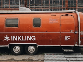 A vintage Airstream trailer with an orange exterior is parked in front of an industrial building with large grid-like windows. The trailer features the word 'INKLING' in bold white letters and some additional text, suggesting it is associated with a brand or company. A website URL is also visible on the trailer.