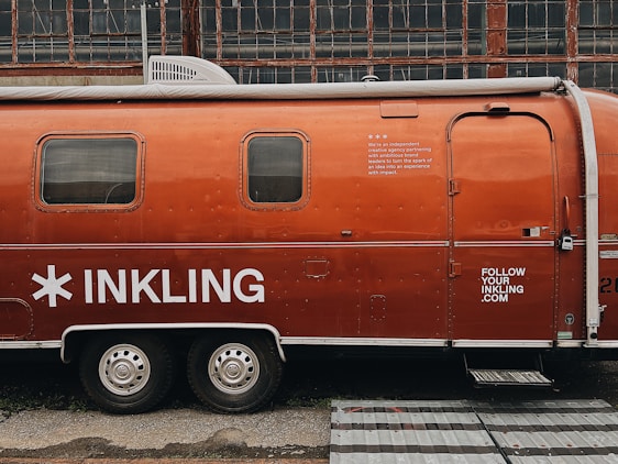 A vintage Airstream trailer with an orange exterior is parked in front of an industrial building with large grid-like windows. The trailer features the word 'INKLING' in bold white letters and some additional text, suggesting it is associated with a brand or company. A website URL is also visible on the trailer.