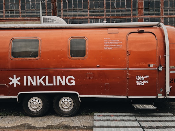 A vintage Airstream trailer with an orange exterior is parked in front of an industrial building with large grid-like windows. The trailer features the word 'INKLING' in bold white letters and some additional text, suggesting it is associated with a brand or company. A website URL is also visible on the trailer.