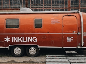 A vintage Airstream trailer with an orange exterior is parked in front of an industrial building with large grid-like windows. The trailer features the word 'INKLING' in bold white letters and some additional text, suggesting it is associated with a brand or company. A website URL is also visible on the trailer.