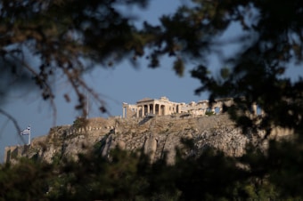 An ancient temple structure is seen atop a rugged hill surrounded by trees in the foreground. The Greek flag flutters on a pole at the left side of the hill, and the clear blue sky forms the backdrop to the scene.