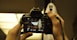 A person is holding a Nikon camera, focusing on a ghostly figure with a white sheet and black eye and mouth cutouts in a dimly lit environment. The camera screen displays the ghost image with camera settings visible.