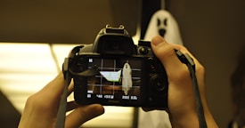 A person is holding a Nikon camera, focusing on a ghostly figure with a white sheet and black eye and mouth cutouts in a dimly lit environment. The camera screen displays the ghost image with camera settings visible.
