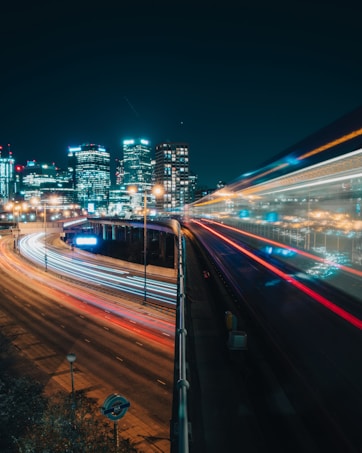 Cityscape at night showcasing a vibrant skyline with illuminated office buildings. Long exposure photography captures light trails of vehicles on a multi-lane highway, creating dynamic streaks of red, white, and blue, indicating motion and energy.