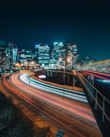 A dynamic urban landscape featuring a night cityscape with skyscrapers illuminated by vibrant lights. Long exposure captures of traffic on a highway create colorful light trails, adding a sense of motion and energy to the scene.
