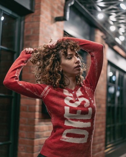 A person with curly hair is standing outdoors in an urban setting, wearing a red top with white lettering. The background shows a brick wall and nighttime lighting with decorative elements.