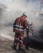 Two construction workers wearing reflective orange suits and white helmets are working on a road at night. One is holding a shovel while the other is engaged in raking or smoothing the asphalt. The scene is enveloped in a cloud of dust and smoke from the construction work.