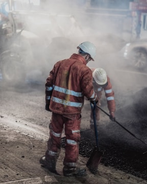 Two construction workers wearing reflective orange suits and white helmets are working on a road at night. One is holding a shovel while the other is engaged in raking or smoothing the asphalt. The scene is enveloped in a cloud of dust and smoke from the construction work.