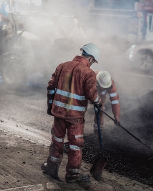 Two construction workers wearing reflective orange suits and white helmets are working on a road at night. One is holding a shovel while the other is engaged in raking or smoothing the asphalt. The scene is enveloped in a cloud of dust and smoke from the construction work.