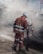 Two construction workers wearing reflective orange suits and white helmets are working on a road at night. One is holding a shovel while the other is engaged in raking or smoothing the asphalt. The scene is enveloped in a cloud of dust and smoke from the construction work.