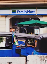 A street scene depicting the entrance to a FamilyMart convenience store with two blue tuk-tuks parked in front. The store has a green awning and signage in English and another language. A person is partially visible inside one of the tuk-tuks.