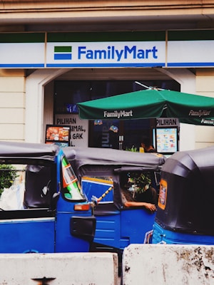 A street scene depicting the entrance to a FamilyMart convenience store with two blue tuk-tuks parked in front. The store has a green awning and signage in English and another language. A person is partially visible inside one of the tuk-tuks.