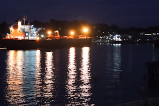 Nighttime shot of a ship illuminated by deck lights, reflecting on the water.