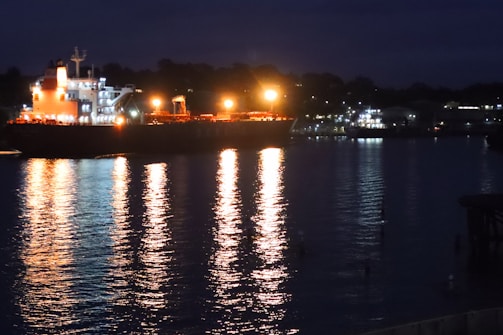 Nighttime shot of a ship illuminated by deck lights, reflecting on the water.