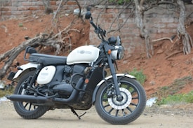 A classic style motorcycle with a white and black color scheme is parked on a dirt path. The background features an old, weathered brick wall and twisted tree roots. The motorcycle has a vintage aesthetic with round headlights and a leather seat.