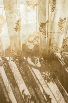 Delicate lace curtains with floral patterns hang in front of a window, casting intricate, ornate shadows across a wooden floor. The sunlight filters through the fabric, creating a serene and ethereal atmosphere.