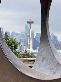 A cityscape featuring a prominent futuristic tower, surrounded by modern skyscrapers and framed by a large circular metal structure. The cloudy sky casts a muted, grayish tone over the urban scene.