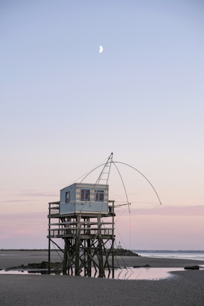 A small wooden cabin stands elevated on stilts above a sandy beach with shallow water reflecting its structure. The cabin has a fishing net apparatus extending from its side, and calm waves lap gently at the shore. In the background, the horizon is bathed in soft pastel colors as the sun sets, and a crescent moon is visible in the twilight sky.
