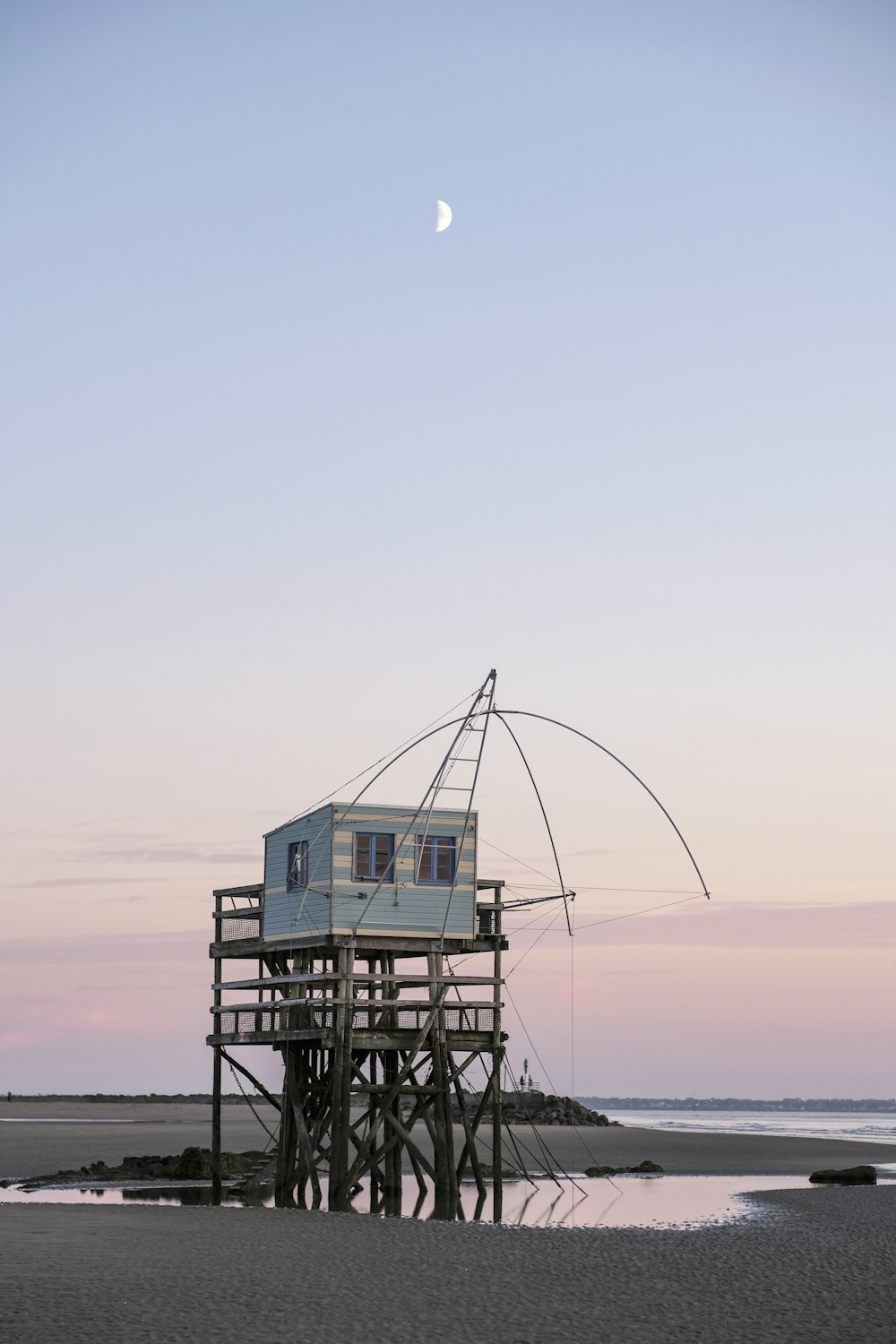 a house on stilts on the beach with a half moon in the sky