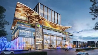 A modern building with a striking architectural design, featuring glass facades and large, decorative overhangs. Illuminated signs for various luxury brands are visible on the exterior. Bright, colorful lights and fountains add vibrancy to the street level, with a few trees and lampposts lining the road.