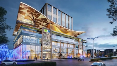 A modern building with a striking architectural design, featuring glass facades and large, decorative overhangs. Illuminated signs for various luxury brands are visible on the exterior. Bright, colorful lights and fountains add vibrancy to the street level, with a few trees and lampposts lining the road.