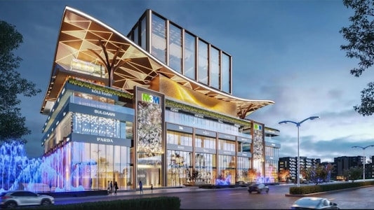 A modern building with a striking architectural design, featuring glass facades and large, decorative overhangs. Illuminated signs for various luxury brands are visible on the exterior. Bright, colorful lights and fountains add vibrancy to the street level, with a few trees and lampposts lining the road.