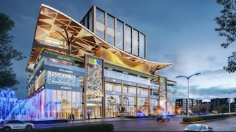 A modern building with a striking architectural design, featuring glass facades and large, decorative overhangs. Illuminated signs for various luxury brands are visible on the exterior. Bright, colorful lights and fountains add vibrancy to the street level, with a few trees and lampposts lining the road.