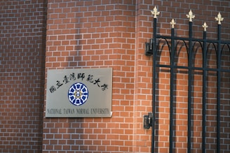 A sign for National Taiwan Normal University is mounted on a red brick wall next to a decorative black metal fence with golden finials.