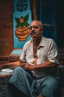 An elderly man with a contemplative expression sits outside, wearing a light-colored shirt and a checkered cloth. Behind him is a blue wall featuring a painted symbol and a decorative image of a pot with leaves.