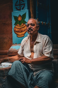 An elderly man with a contemplative expression sits outside, wearing a light-colored shirt and a checkered cloth. Behind him is a blue wall featuring a painted symbol and a decorative image of a pot with leaves.