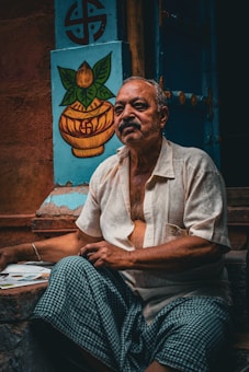 An elderly man with a contemplative expression sits outside, wearing a light-colored shirt and a checkered cloth. Behind him is a blue wall featuring a painted symbol and a decorative image of a pot with leaves.