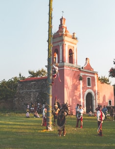 A group of individuals dressed in traditional attire are gathered around a tall pole in front of an old pink church. One person is climbing the pole while others, wearing vibrant costumes with feathers, stand below. A small audience watches the scene on a grassy area.