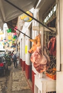 A busy street scene in a rustic market area with a hanging pig carcass displayed outside a butcher shop. Colorful papel picado decorations hang overhead, and pedestrians walk along the narrow sidewalk. Several parked cars line the cobblestone street beside white-walled buildings with red bases.