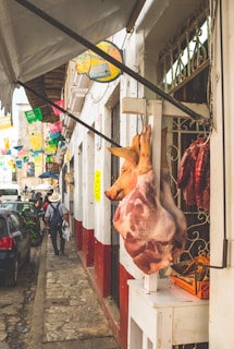 A busy street scene in a rustic market area with a hanging pig carcass displayed outside a butcher shop. Colorful papel picado decorations hang overhead, and pedestrians walk along the narrow sidewalk. Several parked cars line the cobblestone street beside white-walled buildings with red bases.