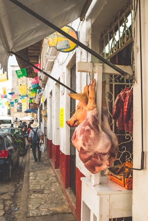 A busy street scene in a rustic market area with a hanging pig carcass displayed outside a butcher shop. Colorful papel picado decorations hang overhead, and pedestrians walk along the narrow sidewalk. Several parked cars line the cobblestone street beside white-walled buildings with red bases.