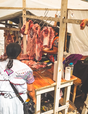 A woman stands at a market stall where fresh cuts of meat are hanging from hooks. The stall is covered with a white canopy, and the woman serving behind the counter is preparing the meat. The scene conveys a typical open-air market setting.