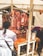 A woman stands at a market stall where fresh cuts of meat are hanging from hooks. The stall is covered with a white canopy, and the woman serving behind the counter is preparing the meat. The scene conveys a typical open-air market setting.