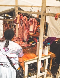 A woman stands at a market stall where fresh cuts of meat are hanging from hooks. The stall is covered with a white canopy, and the woman serving behind the counter is preparing the meat. The scene conveys a typical open-air market setting.