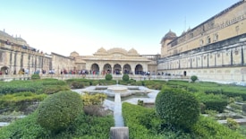 A historic architectural structure features intricate domes and arches, surrounded by well-manicured gardens with lush green hedges and pathways. The area is bustling with numerous visitors, indicating it is a popular tourist attraction. The stone fa&ccedil;ade of the building shows signs of aging, adding to its charm.
